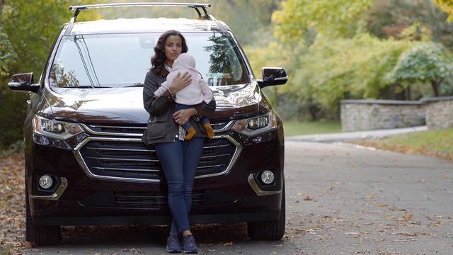 Athena Karkanis in a black jacket and blue jeans holding a baby in front of her car.